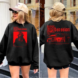 Op de afbeelding: Zwarte sweatshirt met rode grafische tekst en afbeeldingen. De voorkant heeft een rode rechthoek met een silhouet en het woord "Obsession". De achterkant heeft het woord "Obsession" en een huisafbeelding. De sweatshirt wordt door een persoon gedragen.