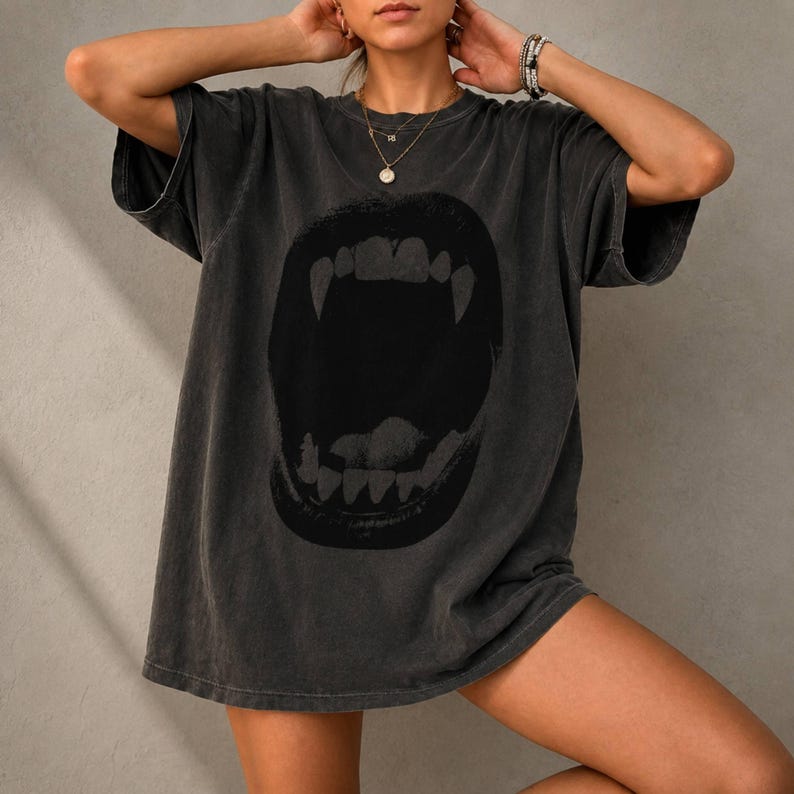 May include: A charcoal gray short-sleeve t-shirt featuring a large, dark graphic print of an open mouth with prominent fangs. The shirt has a relaxed, oversized fit with a crew neckline and a faded, vintage-inspired wash throughout the fabric.