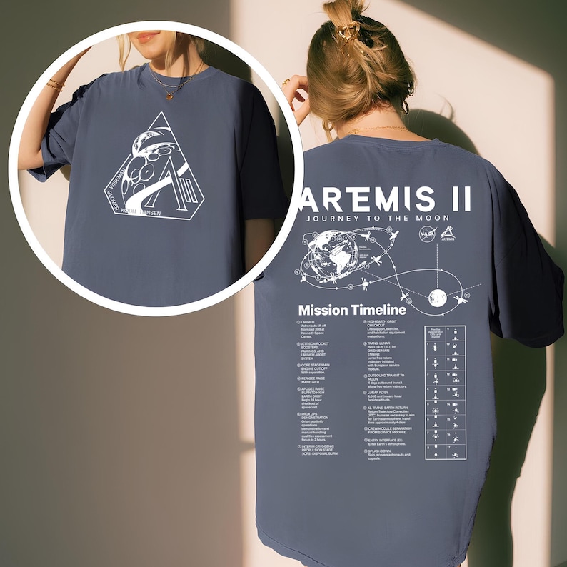 May include: A blue t-shirt featuring a white graphic on the front and back. The front graphic is a triangle with an astronaut and the text "GLOVER, KATHY, HANSEN". The back graphic says "ARTEMIS II JOURNEY TO THE MOON" with a mission timeline.