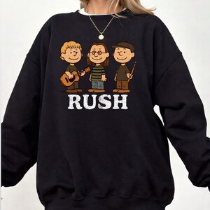 K&ouml;nnte beinhalten: Schwarzes Sweatshirt mit einer Cartoon-Illustration von drei Musikern und dem Wort "RUSH" in wei&szlig;en Blockbuchstaben. Eine Figur spielt Gitarre, eine andere tr&auml;gt eine Brille und die dritte h&auml;lt Trommelst&ouml;cke. Das Design ist eine Hommage an die Band Rush.