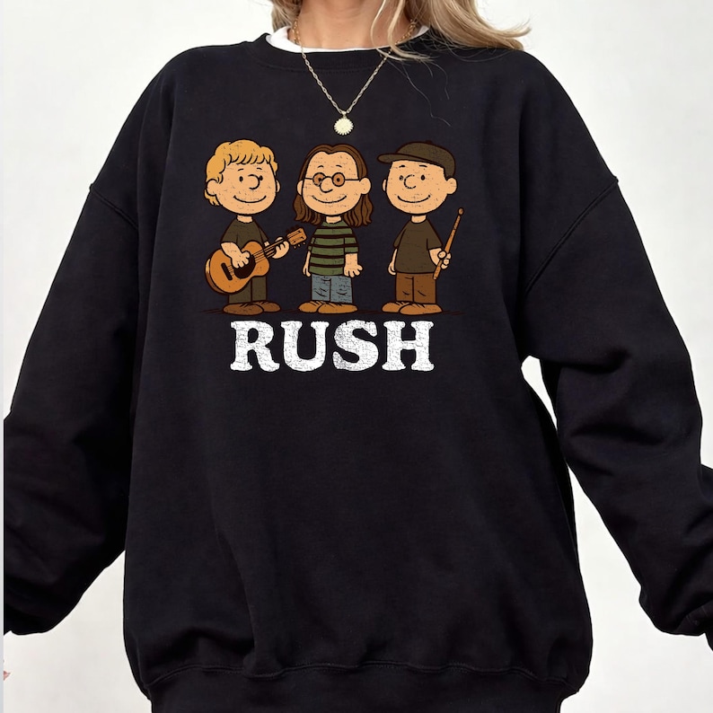 K&ouml;nnte beinhalten: Schwarzes Sweatshirt mit einer Cartoon-Illustration von drei Musikern und dem Wort "RUSH" in wei&szlig;en Blockbuchstaben. Eine Figur spielt Gitarre, eine andere tr&auml;gt eine Brille und die dritte h&auml;lt Trommelst&ouml;cke. Das Design ist eine Hommage an die Band Rush.