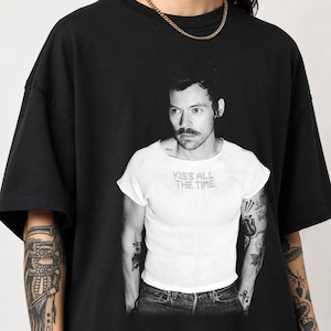 May include: A black short-sleeved t-shirt featuring a large black and white graphic print of a person with a mustache wearing a white shirt with the text KISS ALL THE TIME. The person has visible arm tattoos and wears a gold chain necklace.