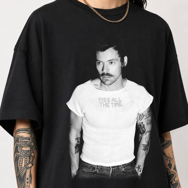 May include: A black short-sleeved t-shirt featuring a large black and white graphic print of a person with a mustache wearing a white shirt with the text KISS ALL THE TIME. The person has visible arm tattoos and wears a gold chain necklace.