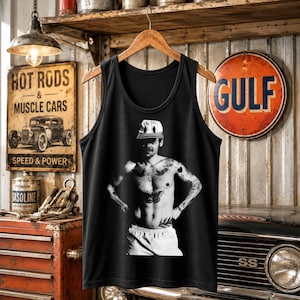 May include: A black sleeveless tank top featuring a centered black and white graphic print of a person wearing a baseball cap, with visible chest and arm tattoos, and wearing light-colored shorts, displayed on a wooden hanger.