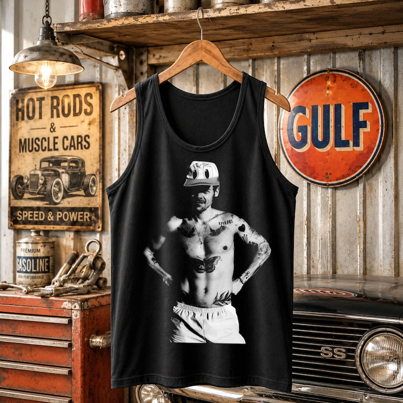 May include: A black sleeveless tank top featuring a centered black and white graphic print of a person wearing a baseball cap, with visible chest and arm tattoos, and wearing light-colored shorts, displayed on a wooden hanger.