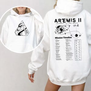May include: White hoodie featuring a black graphic on the front and back. The front graphic is a triangle with a space-themed design and text. The back displays "ARTEMIS II JOURNEY TO THE MOON" with a mission timeline.