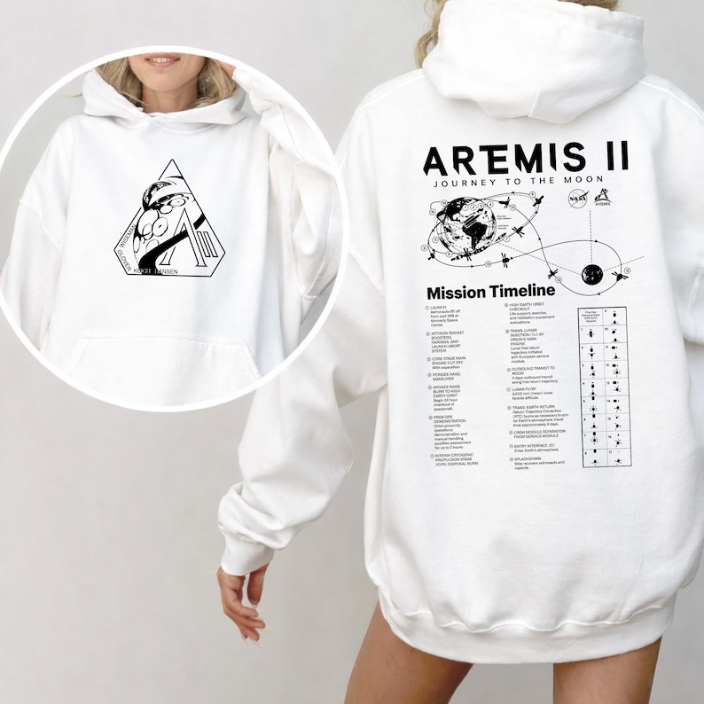 May include: White hoodie featuring a black graphic on the front and back. The front graphic is a triangle with a space-themed design and text. The back displays "ARTEMIS II JOURNEY TO THE MOON" with a mission timeline.