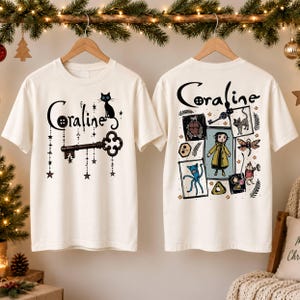May include: Two off-white t-shirts featuring Coraline graphics. The left shirt displays a black cat, a skeleton key, and the text Coraline. The right shirt shows a collage of characters and items from the film, including a girl in a yellow coat.