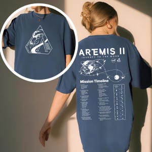 May include: A navy blue t-shirt featuring the Artemis II mission. The front displays a white graphic within a triangle, while the back showcases a mission timeline and the words "ARTEMIS II JOURNEY TO THE MOON".