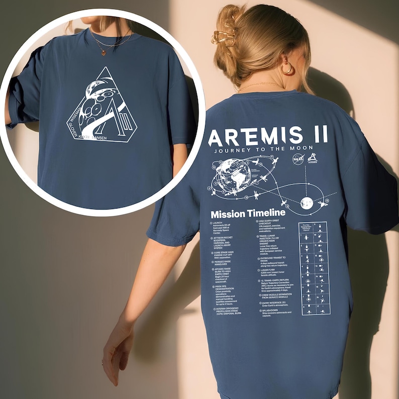 May include: A navy blue t-shirt featuring the Artemis II mission. The front displays a white graphic within a triangle, while the back showcases a mission timeline and the words "ARTEMIS II JOURNEY TO THE MOON".