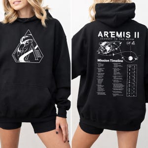 May include: Black hooded sweatshirt with a front graphic featuring a stylized astronaut and the text "Wiseman, Glover, Koch, Hansen". The back of the sweatshirt displays "Artemis II Journey to the Moon" with a mission timeline graphic.