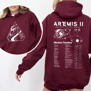 May include: Burgundy hoodie with white graphic on the front and back. The front graphic is a white triangle with a space-themed design and text. The back features the text "ARTEMIS II JOURNEY TO THE MOON" and a mission timeline.