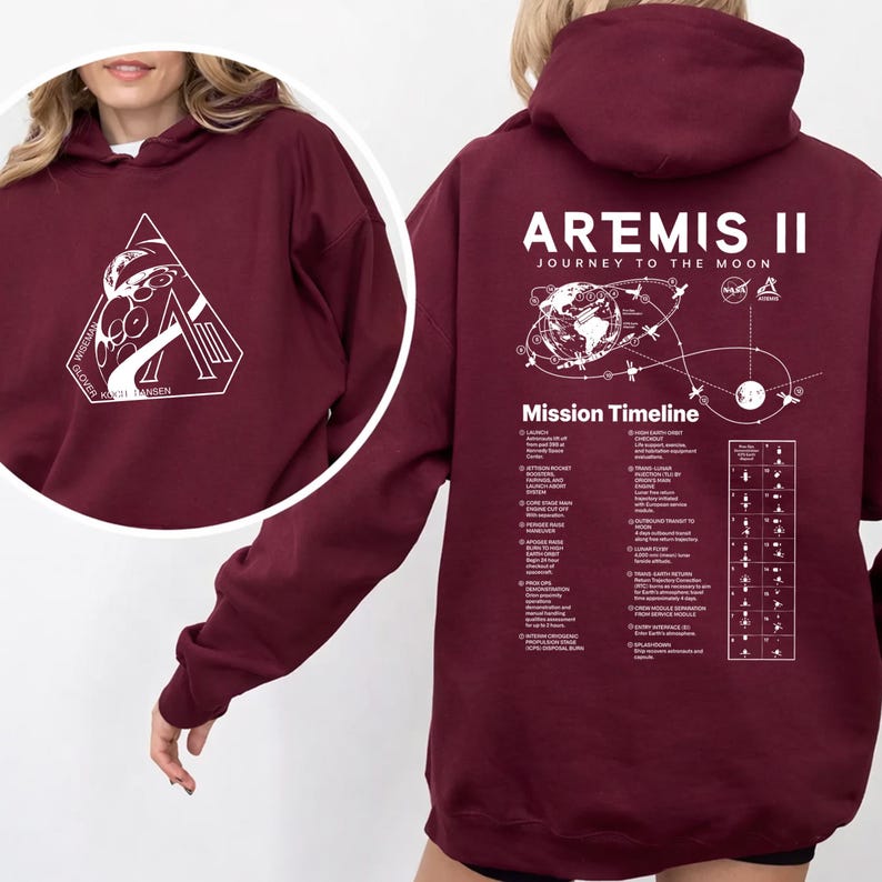 May include: Burgundy hoodie with white graphic on the front and back. The front graphic is a white triangle with a space-themed design and text. The back features the text "ARTEMIS II JOURNEY TO THE MOON" and a mission timeline.