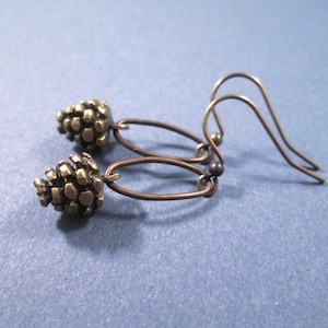 May include: A pair of bronze-toned metal earrings featuring small, textured pinecone charms suspended from simple oval-shaped wire loops. The earrings include curved ear wires for attachment, showcasing a dark metallic finish throughout the design.