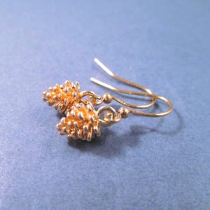 May include: A pair of gold-toned earrings featuring small, textured pinecone-shaped charms suspended from curved ear wires. The metallic finish reflects light, highlighting the intricate, layered scales of the pinecone design on each earring.