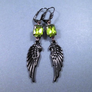 May include: A pair of dangle earrings featuring a dark gunmetal finish. Each earring has a bright green, rectangular gemstone and a detailed wing charm. The earrings are suspended from lever-back closures.