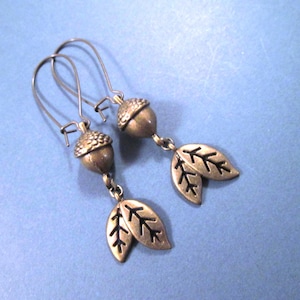 May include: A pair of antique bronze-toned earrings. Each earring features an acorn charm with a textured cap and a pair of leaf charms. The earrings hang from curved ear wires, set against a blue background.