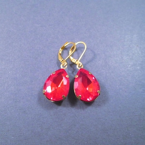May include: A pair of gold-tone leverback earrings featuring faceted, pear-shaped red crystals. Each crystal is secured in a gold-colored metal setting, showcasing a vibrant, translucent red hue with a polished, reflective surface finish.