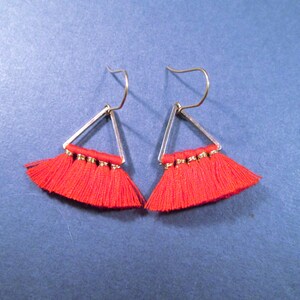 May include: A pair of red tassel earrings. Each earring features a silver-colored triangular frame with a fringe of red tassels. The earrings have silver-colored ear wires. The earrings are displayed against a blue background.