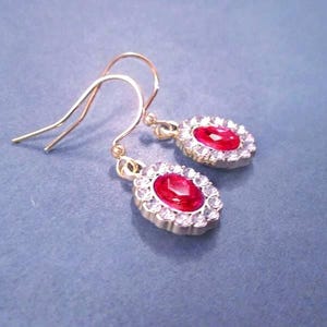 May include: Gold-tone earrings with red oval-shaped center stones surrounded by small clear crystals. The earrings have a classic design with a hook closure. The background is a solid blue-gray color.