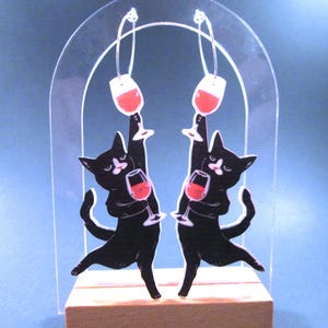 May include: A pair of novelty earrings featuring black cat silhouettes holding wine glasses filled with red liquid. The earrings are suspended from silver hoops and set against a clear acrylic arch and wooden base.