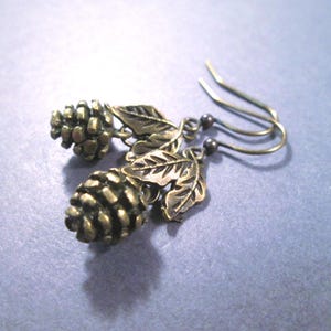 May include: A pair of dangle earrings featuring antique bronze-toned metal charms shaped like pinecones and leaves. Each earring is suspended from a curved metal ear wire, showcasing detailed textures on the pinecone scales and leaf veins.