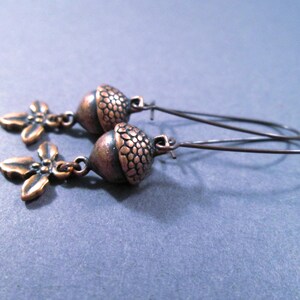 May include: A pair of copper-toned earrings. Each earring features a detailed acorn pendant with a textured cap and a small, four-leaf clover charm. The earrings hang from thin, curved ear wires, creating a nature-inspired accessory.