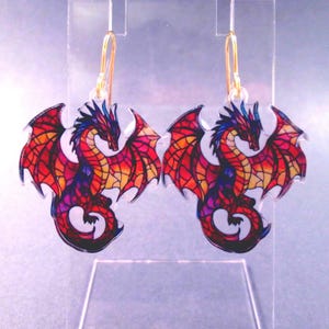 May include: A pair of dragon-shaped earrings. Each earring features a colorful dragon design with red, orange, yellow, and purple hues. The dragon has outstretched wings and is suspended from a gold-colored hook.