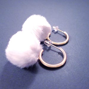 May include: A pair of gold-colored hoop earrings with white, fluffy pom-poms. The earrings are displayed on a dark blue surface, with the focus on the earrings' design and texture. The earrings have a post-back closure.