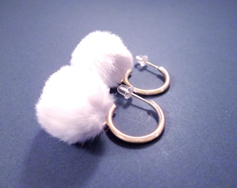 May include: A pair of gold-colored hoop earrings with white, fluffy pom-poms. The earrings are displayed on a dark blue surface, with the focus on the earrings' design and texture. The earrings have a post-back closure.