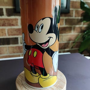 May include: A brown ceramic mug with a cartoon image of Mickey Mouse. Mickey is wearing red shorts and yellow shoes. The mug is sitting on a wooden base.