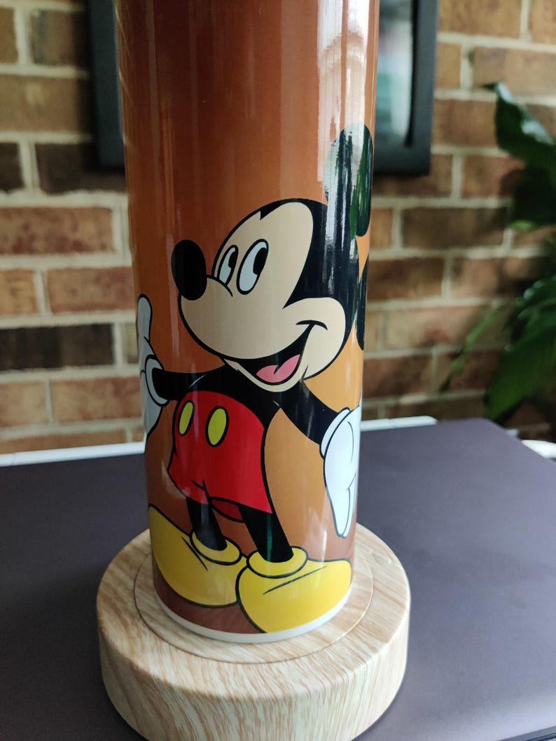 May include: A brown ceramic mug with a cartoon image of Mickey Mouse. Mickey is wearing red shorts and yellow shoes. The mug is sitting on a wooden base.