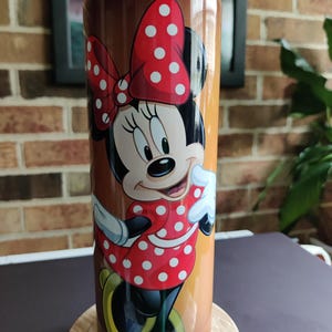 May include: A brown and gold travel mug with a black lid and a colorful image of Minnie Mouse wearing a red dress with white polka dots and a red bow with white polka dots.