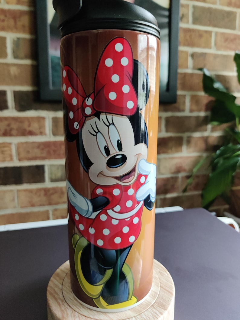 May include: A brown and gold travel mug with a black lid and a colorful image of Minnie Mouse wearing a red dress with white polka dots and a red bow with white polka dots.