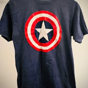 Puede incluir: Una camiseta azul marino con un gr&aacute;fico de escudo de Capit&aacute;n Am&eacute;rica rojo, blanco y azul.