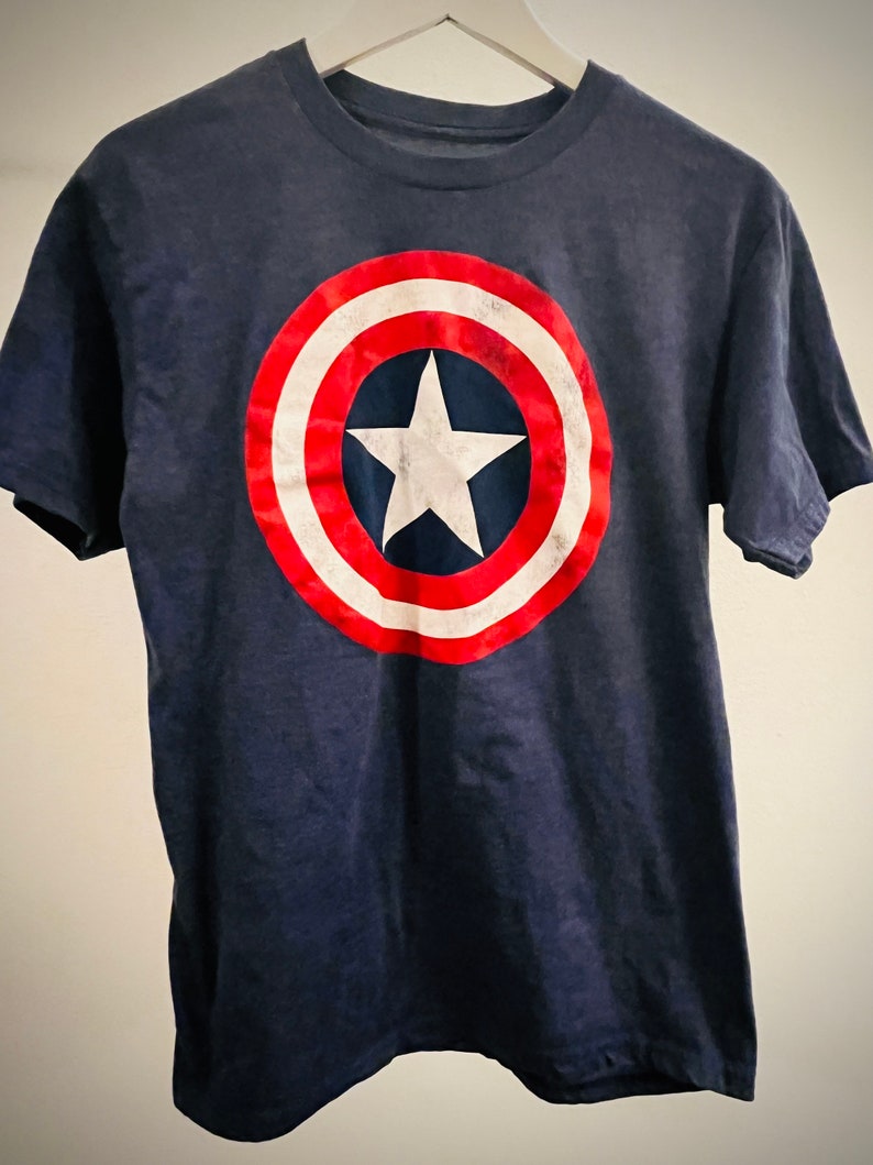 Puede incluir: Una camiseta azul marino con un gr&aacute;fico de escudo de Capit&aacute;n Am&eacute;rica rojo, blanco y azul.