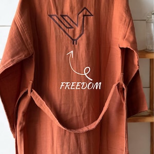 Könnte beinhalten: Rostfarbener Leinen-Bademantel mit geometrischem Vogeldesign auf dem Rücken. Das Wort "FREEDOM" steht unter dem Vogel. Der Bademantel hat einen passenden Gürtel und hängt an einer weiß gefliesten Wand.