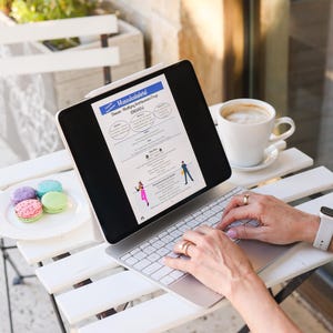 Könnte beinhalten: Eine Person benutzt ein Tablet mit Tastatur auf einem weißen Tisch, mit einer Tasse Kaffee und Makronen. Das Tablet zeigt ein Dokument mit dem Titel "Muskuloskelettal". Die Person trägt Ringe und eine Uhr.