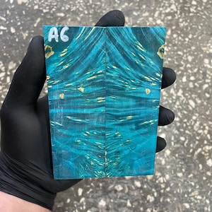 May include: A rectangular piece of wood with a vibrant teal and gold pattern. The wood grain features swirling patterns and knots. The top left corner has the handwritten text "A6". The wood is held by a gloved hand.