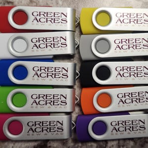 May include: A collection of ten swivel USB flash drives in various colors including red, white, blue, green, pink, yellow, silver, black, orange, and purple. Each drive features a silver metal clip printed with the text GREEN ACRES SEW & VAC.