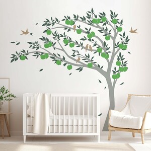 Op de afbeelding: Een wit babybedje met een wit dekentje in een kinderkamer met een grijze en groene boomwanddecoratie met bruine vogels.