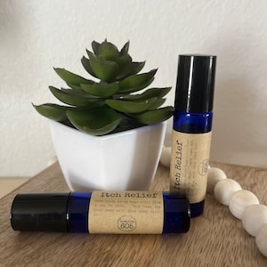 Puede incluir: Dos frascos de vidrio azul con tapas negras. Los frascos están etiquetados con "Itch Relief" y contienen un remedio natural para las picaduras de insectos. El texto de la etiqueta dice: "Cuando esos molestos insectos todavía encuentran una forma de morder, ayuda a aliviar la picazón con este fácil rodillo."