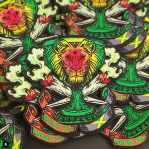 May include: A stack of embroidered patches featuring a green and yellow lion's head inside a green cup with a red and yellow flame on each side. The patches have a green, yellow, and black ribbon with the text "Banda Cup" on it.