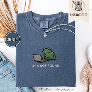 Puede incluir: Una camiseta azul vaquero con un diseño bordado de una rana verde usando un portátil, con el texto "404 NOT FOUND" debajo. También se ve un collar plateado con un colgante. La camiseta está doblada.