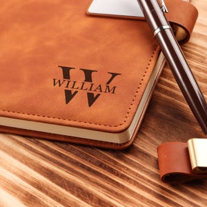 May include: A brown leather journal with the name "WILLIAM" embossed in black. A brown pen rests on the journal, and a leather pen holder is visible. The journal is on a wooden surface.