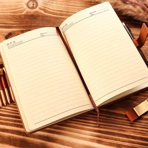 May include: An open, lined notebook with a brown leather cover and a matching pen holder. A small leather case with golf tees is attached to the notebook. A gold-colored metal bookmark rests on the wooden surface.