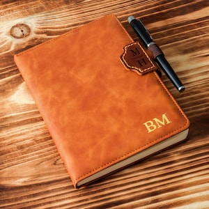 Pode incluir: Um diário de couro castanho com as iniciais "BM" em relevo na capa. Uma caneta repousa sobre o diário, e uma aba de couro com as iniciais "MH" está presa. O diário está sobre uma superfície de madeira.