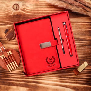 May include: A red leather journal with a silver clasp, a pen, and a pen refill are displayed in a red box. The journal is embossed with the letter "S" and the name "SARAH". The set is on a wooden surface.