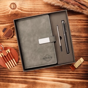 May include: A gray leather-bound journal with a silver clasp and the initials "D.S." is presented in a gift box, alongside a black pen and a pen refill. A golf tee holder and a leather accessory are also visible.