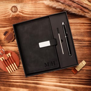 May include: A black leather journal with the initials "MH" engraved on the cover, presented in a gift box with a pen and a pen refill. The journal has a silver clasp. A golf tee holder and other accessories are also visible.